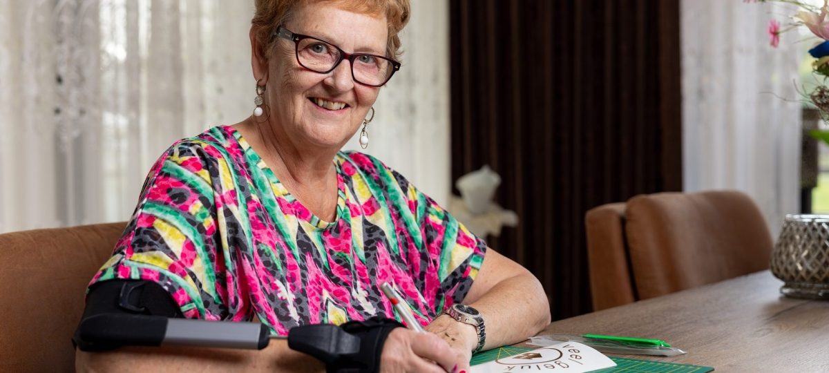 Vrouw aan tafel met STIL brace om arm. Met de arm waar de brace om zit snijdt ze iets uit op papier. Ze lacht in de camera.