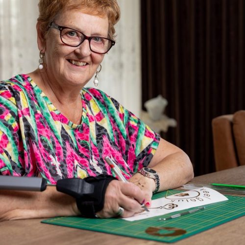 Vrouw aan tafel met STIL brace om arm. Met de arm waar de brace om zit snijdt ze iets uit op papier. Ze lacht in de camera.