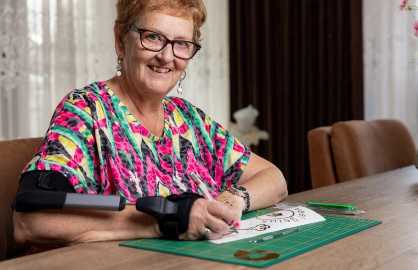 Vrouw aan tafel met STIL brace om arm. Met de arm waar de brace om zit snijdt ze iets uit op papier. Ze lacht in de camera.